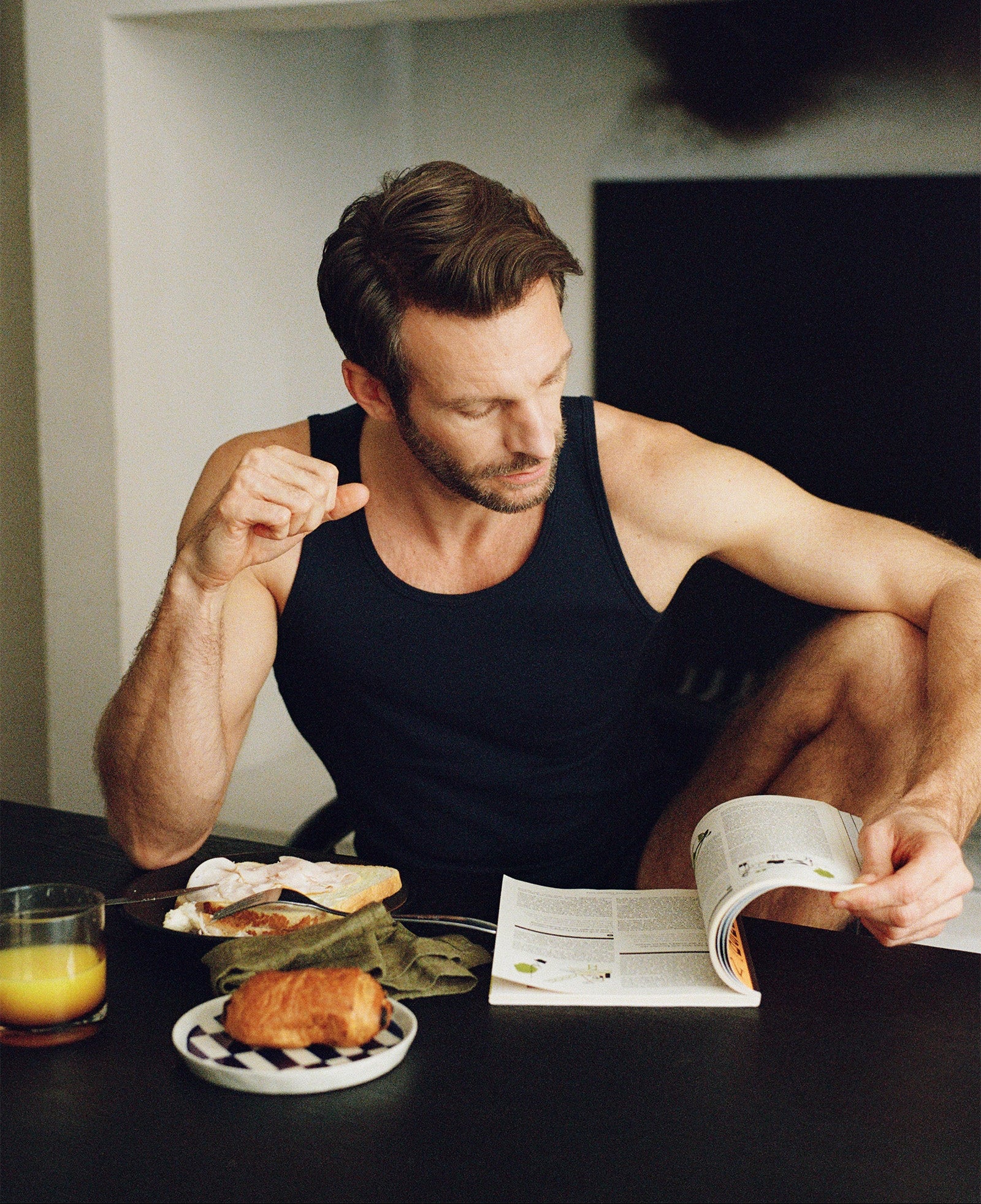 Man sitting at a table in a navy tank top with food and a magazine, looking at the magazine.